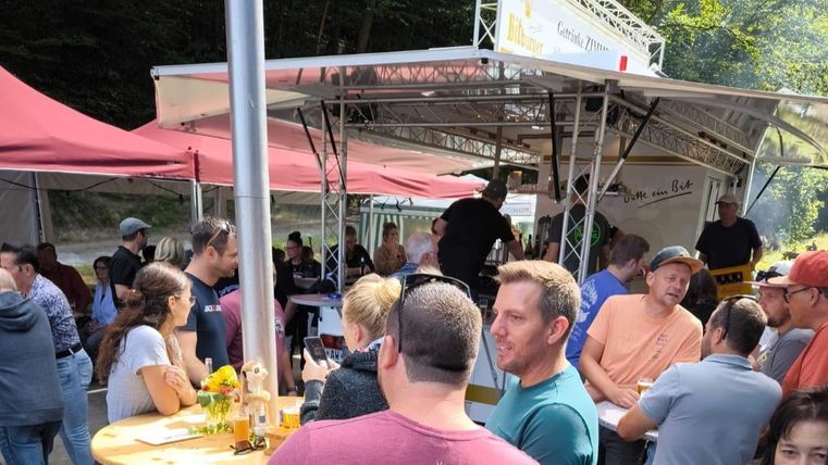 Eine gesellige Veranstaltung mit vielen Menschen, die an einem Essensstand stehen. Es gibt Stühle, Tische und Sonnenschirme im Freien.