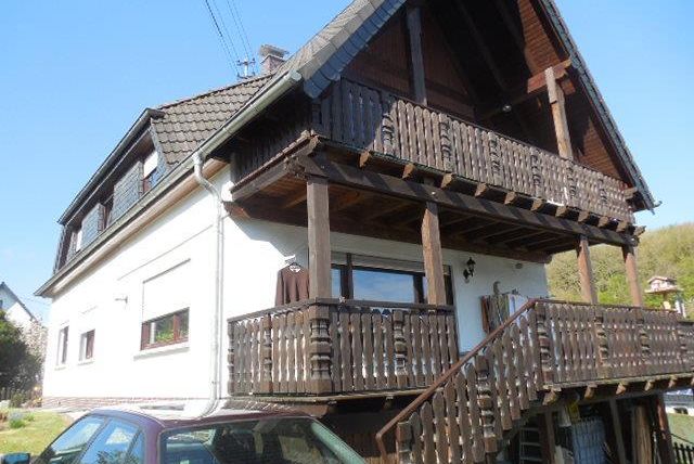 Une maison blanche avec un balcon en bois et un toit en pente. Au premier plan, il y a une voiture et l'environnement est vert.