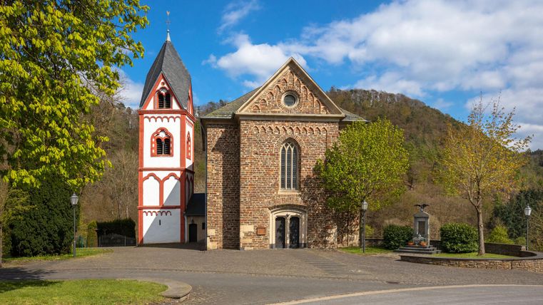 Eine malerische Kirche mit einem roten und weißen Turm liegt eingebettet in eine grüne Landschaft. Im Hintergrund sind sanfte Hügel und ein blauer Himmel zu sehen.