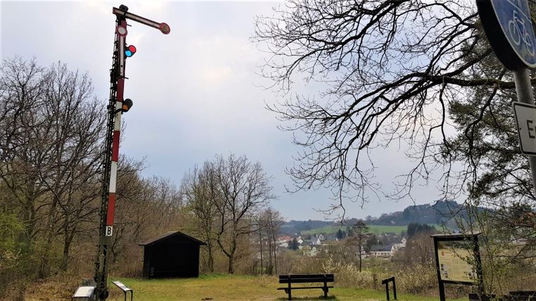 Een spoorwegovergang met een signaal en een bank in een landelijke omgeving. Op de achtergrond zijn bomen en heuvels te zien.