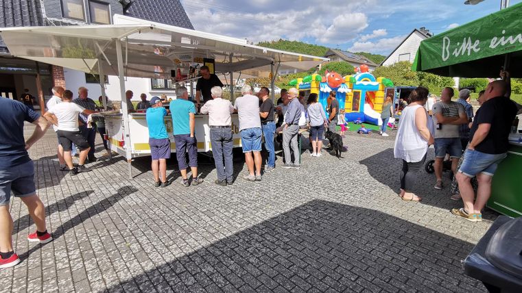 Ein belebter Markt mit vielen Menschen, die an einem Imbissstand warten. Im Hintergrund sind Spielmöglichkeiten für Kinder sowie eine einladende Atmosphäre zu sehen.