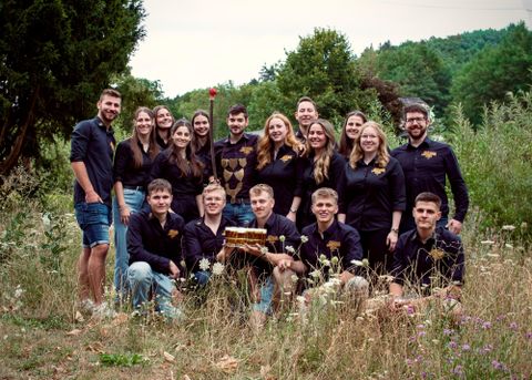 Eine Gruppe von jungen Menschen steht lächelnd auf einer Wiese. Sie machen Daumen hoch und präsentieren eine Partie Getränke auf einem Tablett.