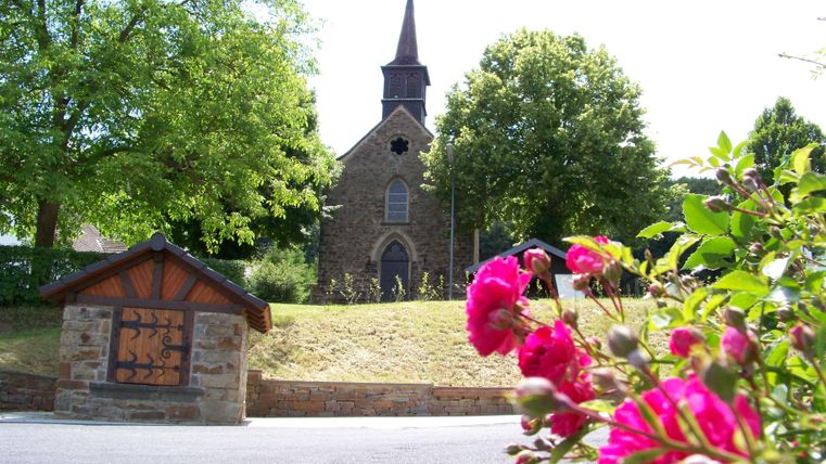Eine kleine Kirche umgeben von Bäumen und bunten Blumen. Der Himmel ist klar und sonnig.