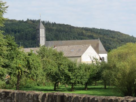 Eine alte Kirche mit einem hohe Turm steht vor einer bewaldeten Hügelkulisse. Im Vordergrund wachsen Obstbäume in einer grünen Wiese.