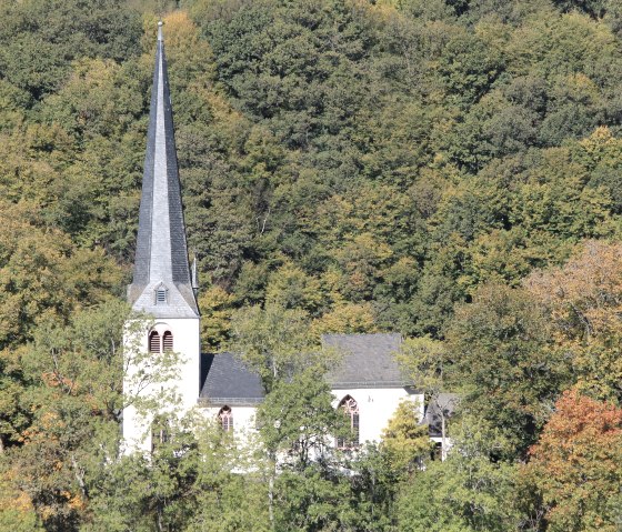 Eine Kapelle mit spitzem Turm steht vor einem dichten, herbstlich gefärbten Wald. Die Bäume zeigen grüne und orangefarbene Blätter., © Walter Schmitz