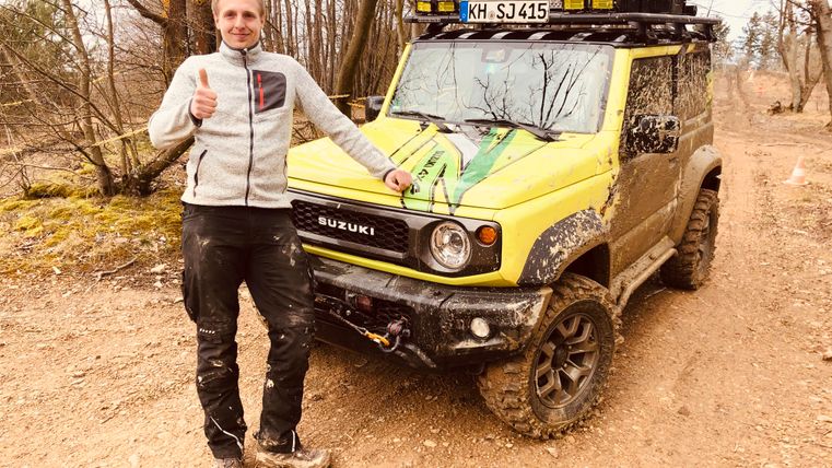 Ein Mann steht neben einem gelben Suzuki Jimny in einem Waldgebiet. Er zeigt mit Daumen nach oben und das Auto ist schmutzig von Offroad-Fahren.