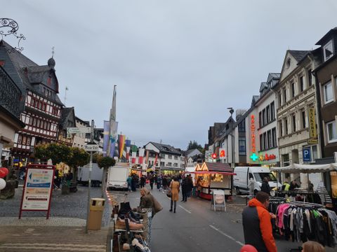 Ein belebter Marktplatz mit Ständen und Menschen. Die Gebäude sind bunt, und es hängt eine Reihe von Flaggen in der Luft.