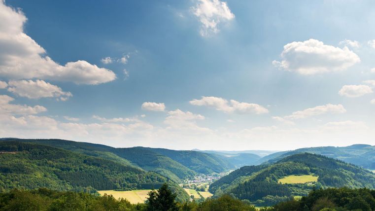 Eine malerische Landschaft mit sanften Hügeln und grünen Wäldern. Der Himmel ist blau und mit vereinzelten Wolken geschmückt.