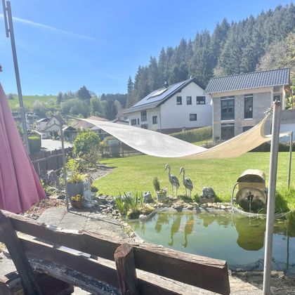 A beautiful garden with a pond and decorative figures. In the background, houses and forests can be seen under bright sunshine.