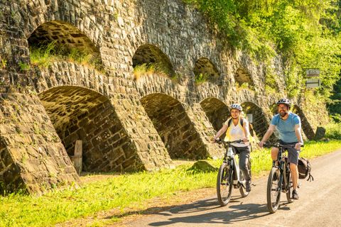 Two cyclists ride along a narrow path beside a stone wall with arches. The surroundings are green and sunny, creating a pleasant atmosphere.