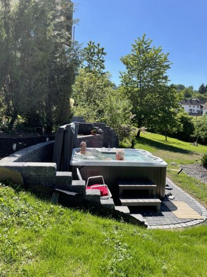 A hot tub outdoors, surrounded by greenery and trees. The sky is blue and the sun is shining.