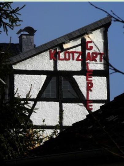 Een historisch vakwerkhuis met het opschrift "KLOTZART GALERIE". De lucht is helder en blauw, en het huis wordt omringd door bomen.