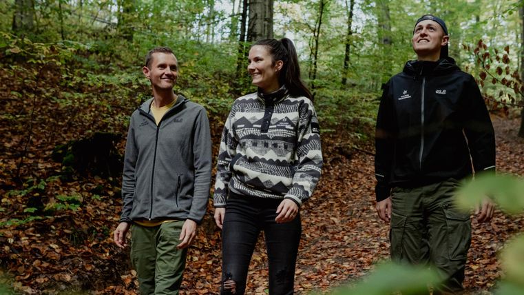 Drei Personen wandern durch einen grünen Wald mit herbstlichen Blättern. Sie lachen und genießen die Natur.