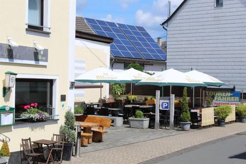 Ein gemütliches Café mit Sonnenschirmen und Tischen im Freien. Im Hintergrund sind Solarpanels auf einem Dach sichtbar.