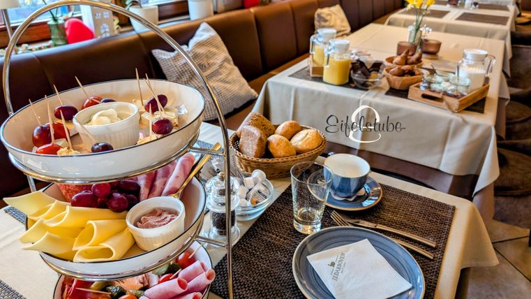Ein stilvoll gedeckter Tisch mit einem dreistöckigen Obst- und Käsebonnet sowie frischen Brötchen. Dazu Getränke und Geschirr für ein gemütliches Frühstück.