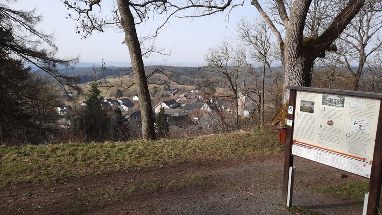Eine Aussicht auf ein kleines Dorf, umgeben von Bäumen und Hügeln. Im Vordergrund steht ein Informationstafel.