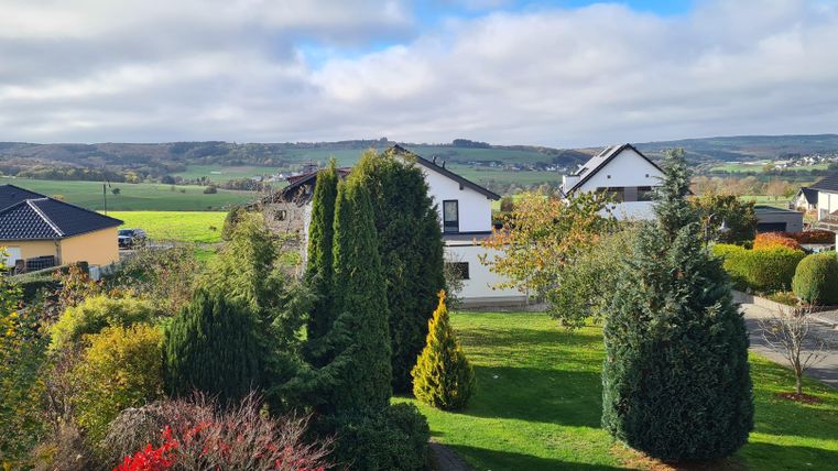 A peaceful rural landscape with a beautiful garden and several houses. The sky is overcast and there are gentle hills in the distance.