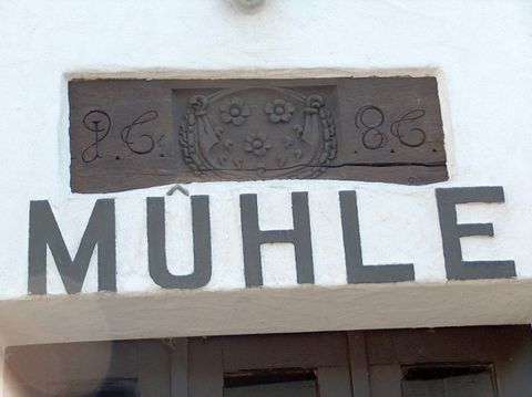 Ein historisches Mühlenschild mit einer eingravierten Darstellung und dem Wort „Mühle“. Es zeigt ein einfaches, rustikales Design.
