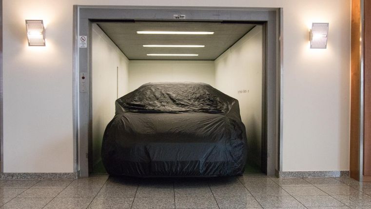 Een auto die met een hoes is bedekt, staat in een lift. De muren zijn licht en de vloer is betegeld.