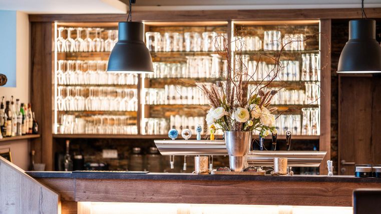 A stylish bar with an appealing wood paneling and a shelf full of glasses. A flower arrangement in a silver vase is in the foreground.