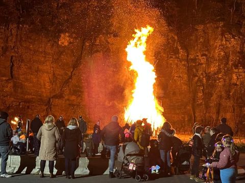 Eine Gruppe von Menschen versammelt sich um ein großes Feuer unter einem Felsen. Die Atmosphäre ist festlich und die Flammen strahlen warmes Licht aus.