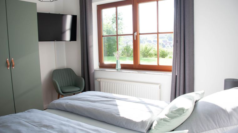 Ein modernes Schlafzimmer mit zwei Betten und einem Fernseher an der Wand. Das Fenster bietet einen Blick ins Grüne und lässt viel Tageslicht hereinfallen.
