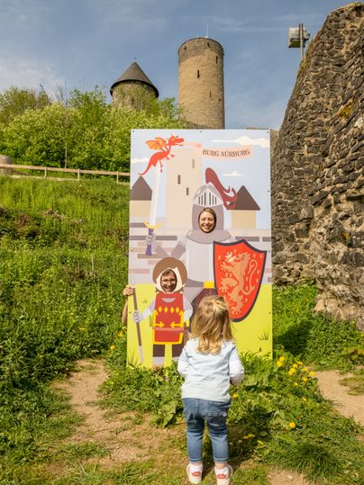 Vor der Burg Nürburg steht eine bunte Fototafel mit Rittermotiv und Ausschnitt für das Gesicht. Ein Kind blickt von hinten zur Tafel, während im Hintergrund die Burgmauern und Türme über grünem Bewuchs aufragen.