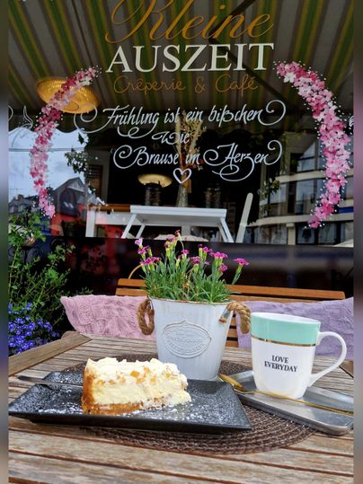 Un morceau de cheesecake sur une assiette noire, une tasse avec l'inscription "LOVE EVERYDAY", et un bouquet de fleurs épanouies sur une table en bois. En arrière-plan, une fenêtre avec l'inscription "Kleine Auszeit" et des fleurs décoratives est visible.