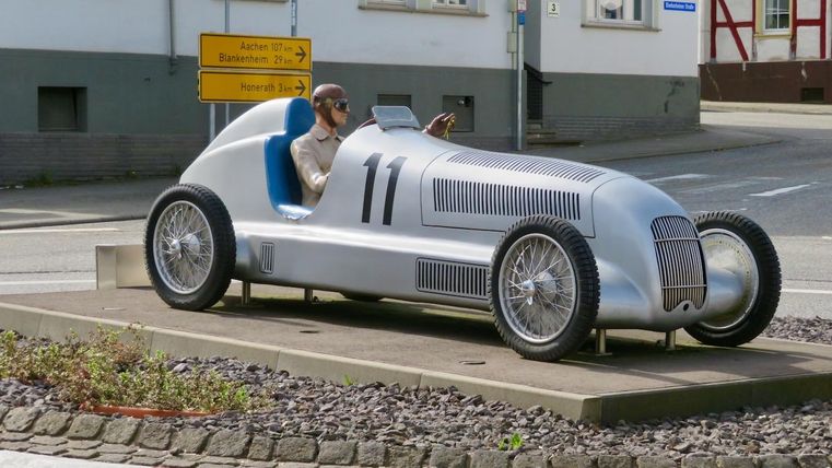 Une voiture de course rétro argentée est stationnée dans un rond-point. À l'intérieur, une figurine qui ressemble à un conducteur est assise, affichant un regard détendu.