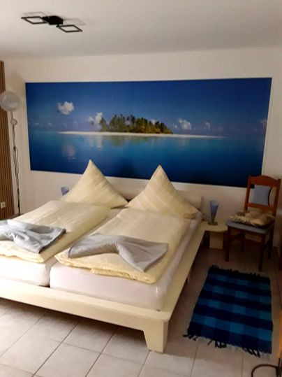 Een gezellige kamer met twee bedden en een grote muurschildering van een rustige strand. Naast de bedden staat een stoel en ligt er een tapijt op de vloer.