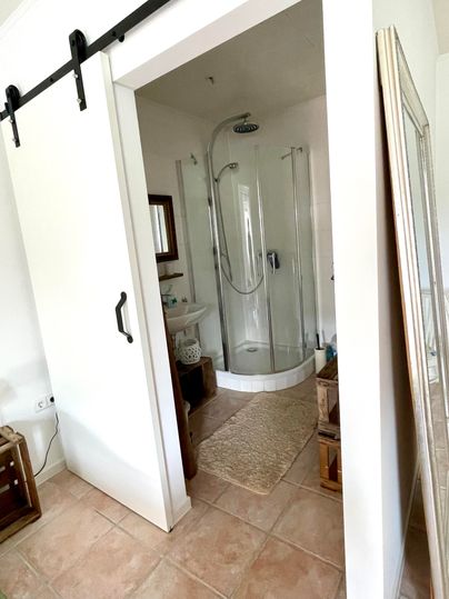 Une salle de bains moderne avec une cabine de douche en verre. Le décor est clair et accueillant, avec un sol en carrelage et un grand miroir.