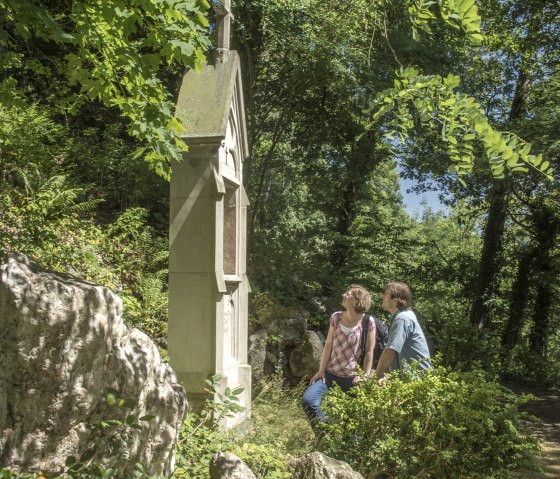 Zwei Personen knien vor einem steinernen Denkmal im Wald auf dem Kreuzweg Adenau. Umgeben von &uuml;ppigem Gr&uuml;n und Felsen., &copy; TI Hocheifel-N&uuml;rburgring, Kappest