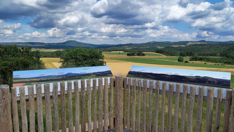 A breathtaking view from an observation platform with lush fields and gentle hills. The sky is adorned with white clouds, providing a clear view of the landscape.
