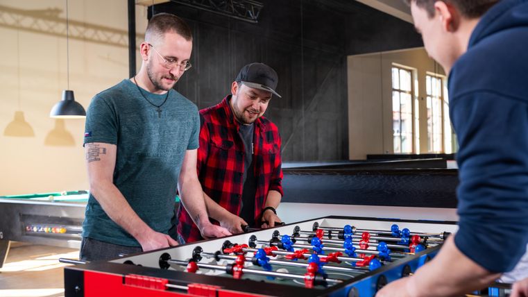 Three men are enthusiastically playing table soccer in a modern room. The atmosphere is relaxed and friendly.