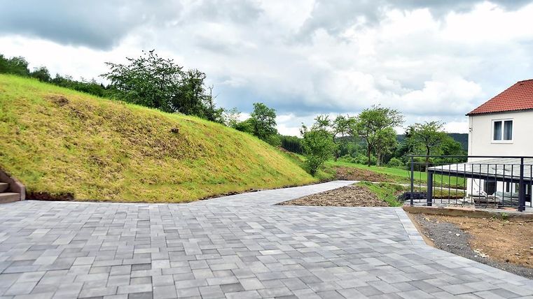 Ein gepflasterter Weg führt zu einem Haus auf einer leicht erhöhten Wiese. Im Hintergrund gibt es Bäume und einen bewölkten Himmel.