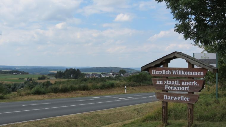 A welcoming sign in the rural setting of Barweiler. In the background, gentle hills and a cloudy sky can be seen.