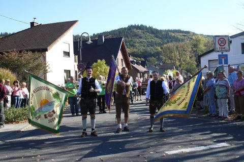 A festive parade featuring people in traditional costumes holding flags. Spectators and a picturesque landscape can be seen in the background.
