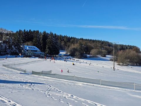 Ein schneebedecktes Feld mit vielen Menschen, die Ski fahren und Spaß haben. Im Hintergrund sind Bäume und ein Gebäude sichtbar.