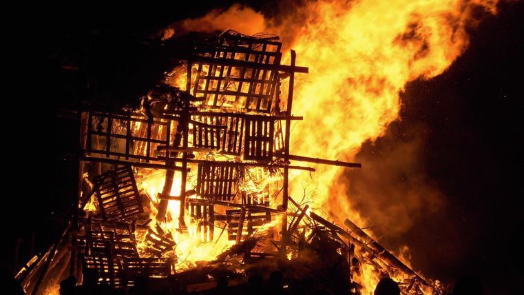 Ein großes Feuer mit brennendem Holz und Flammen. Die Szene ist dunkel, und das Licht des Feuers erhellt die Umgebung.