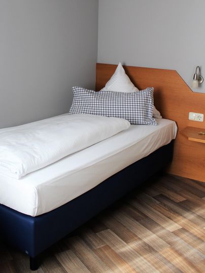 A simple bedroom with a single bed and some pillows. The walls are gray and the floor is wooden.