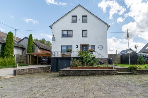 Ein großes, weißes Wohnhaus mit mehreren Fenstern und einem Balkon. Vor dem Haus gibt es einen gepflasterten Hof und einige grüne Pflanzen.