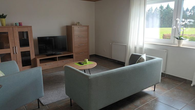 Een moderne woonkamer met twee lichte zetels en een comfortabele bank. De grote ramen laten veel daglicht binnen en bieden uitzicht op het groene landschap.