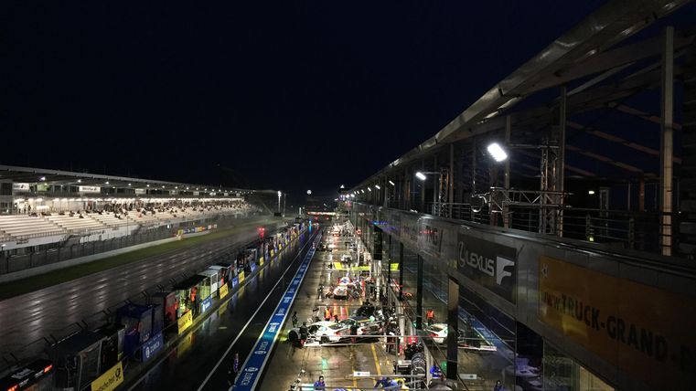 Eine belebte Boxengasse bei Nacht mit mehreren Rennwagen und Lichtern. Die Atmosphäre ist dynamisch und festlich.