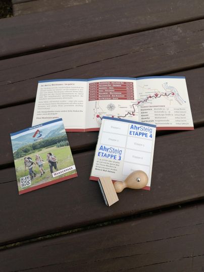Eine Broschüre zeigt eine Wanderroute mit einer Karte und Informationen. Dazu liegt ein Stempel und eine kleine Holzscheibe auf einem Tisch.