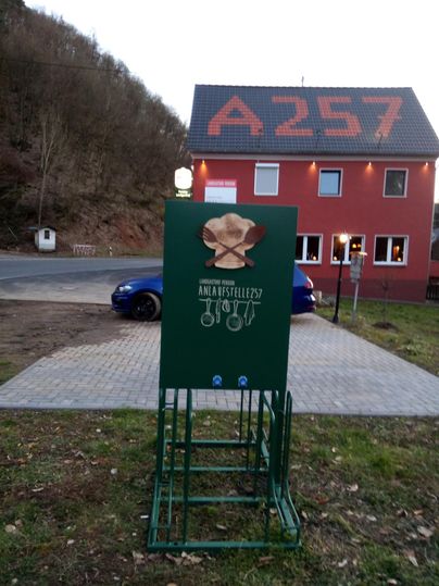 A green sign indicating a rest stop named "ANALSTREIFEN" stands in front of a red building. In the background, a road and a blue car are visible.