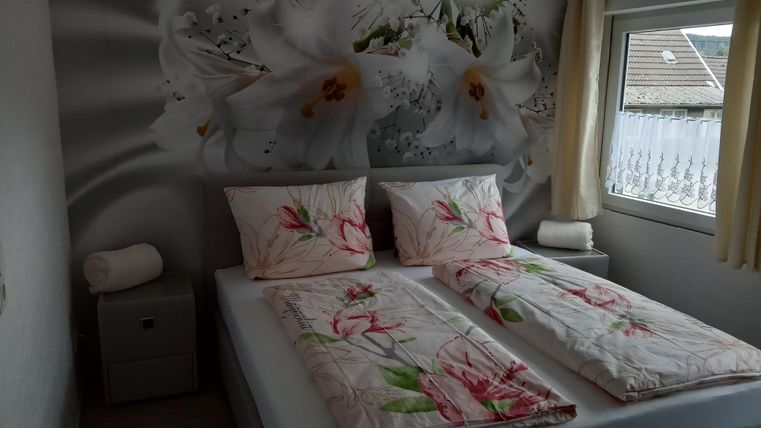 A cozy bedroom with a large bed and floral bedding. On the wall, there is a mural with white flowers, and the room has a window with daylight.