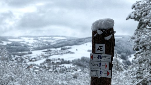 Ein verschneiter Wegweiser im Vordergrund zeigt Wanderwege an. Im Hintergrund erstreckt sich eine winterliche Landschaft mit schneebedeckten H&uuml;geln., &copy; TI Hocheifel-N&uuml;rburgring,S.Schulte
