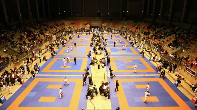 Een sporthal met meerdere wedstrijdvlakken voor competities. Veel toeschouwers en deelnemers zijn aanwezig.
