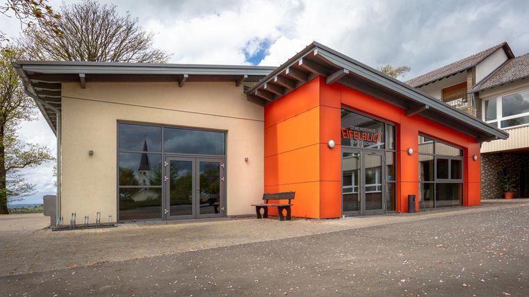 Ein modernes Gebäude mit einer orangefarbenen Fassade. Es gibt große Fenster und eine benachbarte Bank im Außenbereich.