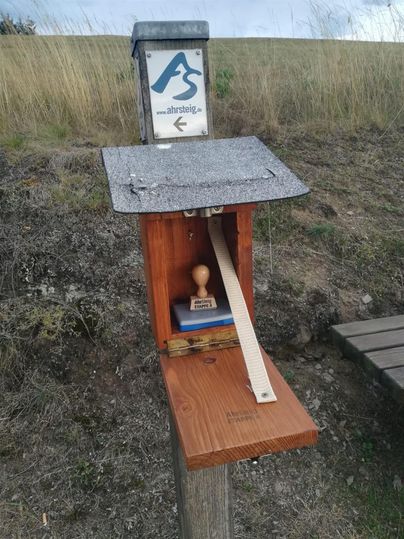 Eine kleine Holzkiste mit einem Stempel und einem langen Holzstück steht an einem Weg. Darüber hängt ein Schild mit einer Webseite und einem Richtungspfeil.